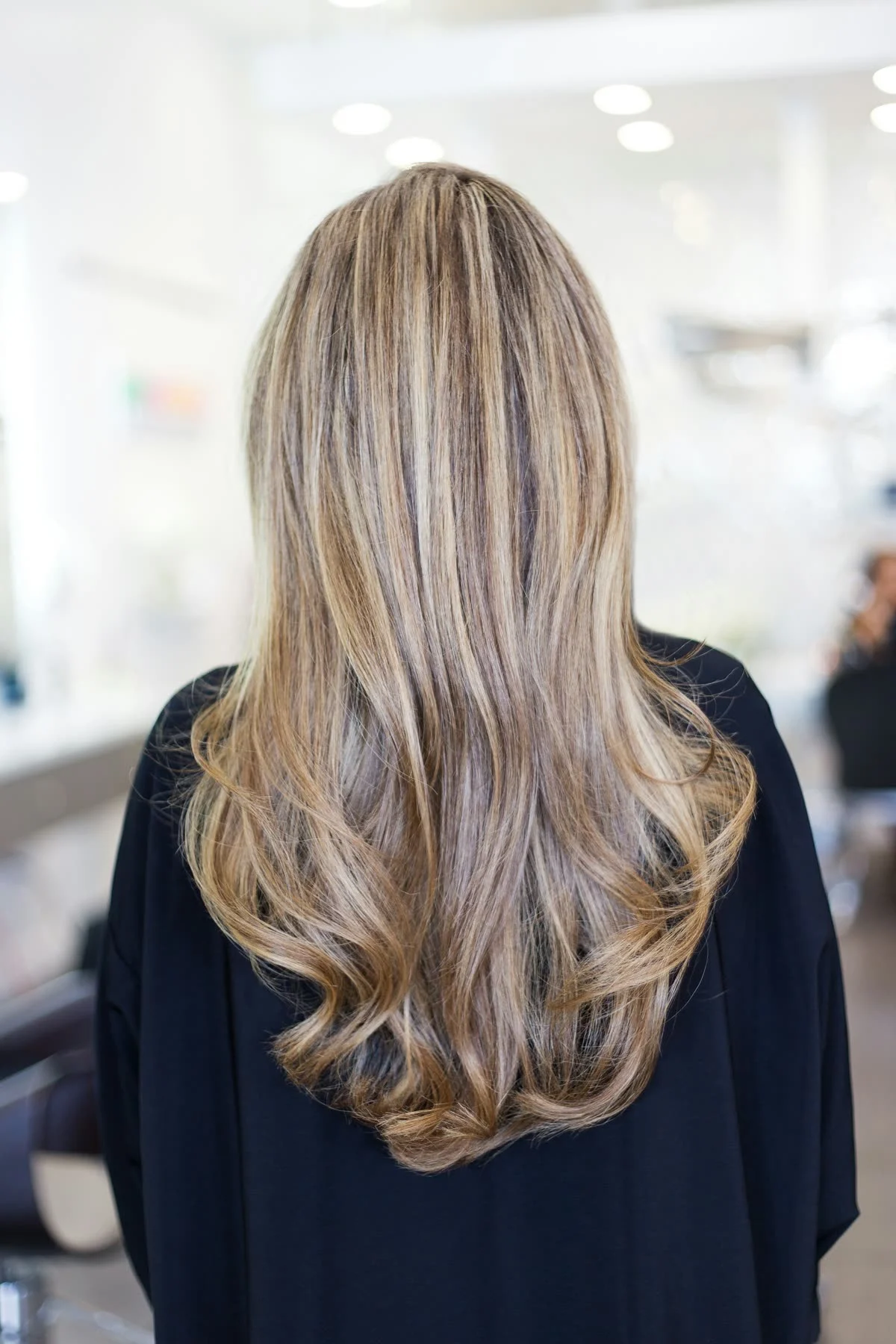A glossy, bouncy blowout in long highlighted hair, seen from behind in a bright salon