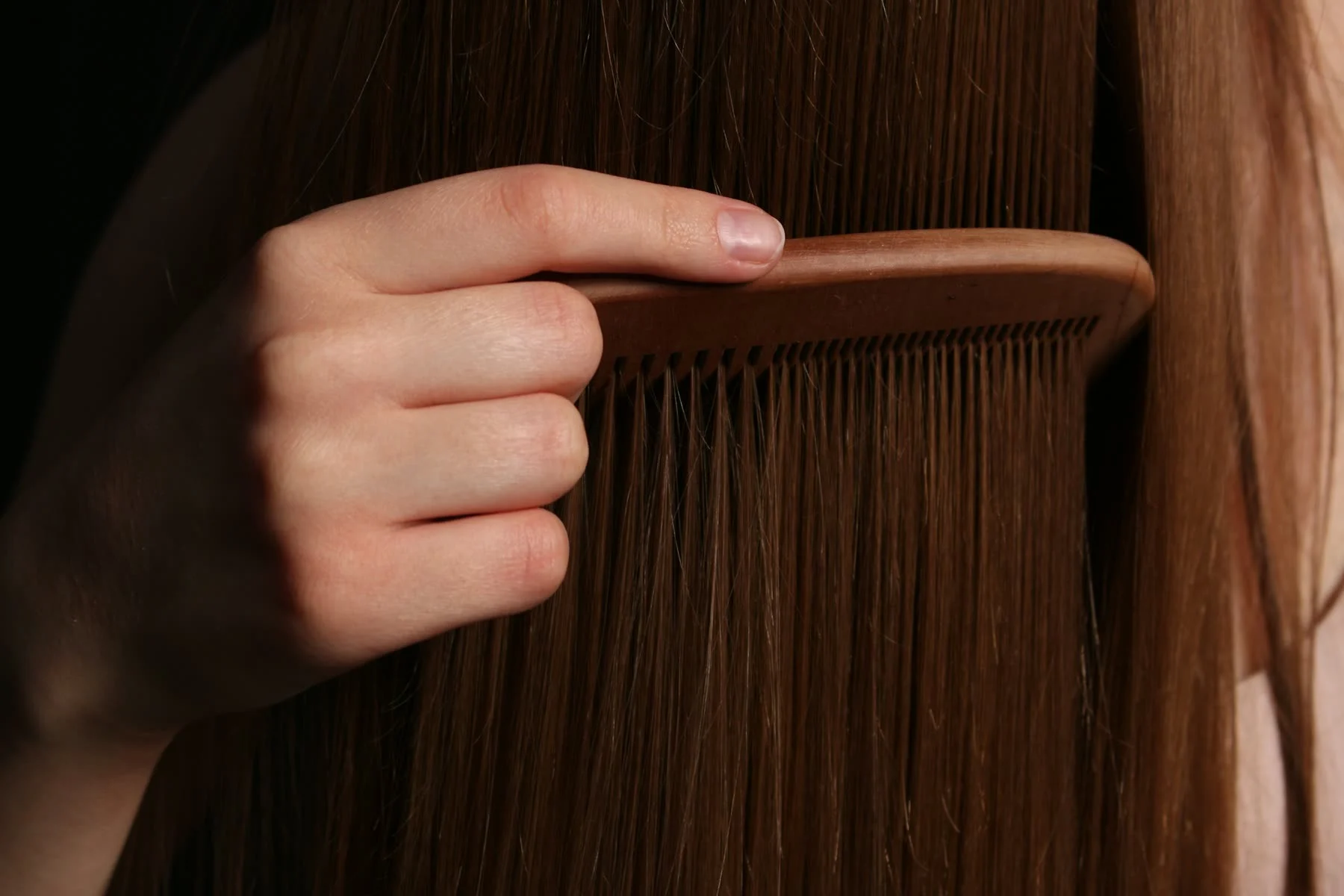A hand draws a wooden comb through long, straight auburn hair